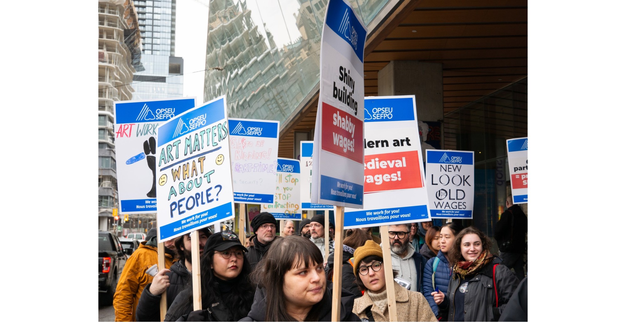 Union members holding signs on sticks picketing outside of the Art Gallery of Ontario after going on strike on March 26. The signs say "New Look, Old Wages" and "Modern art, medieval wages".