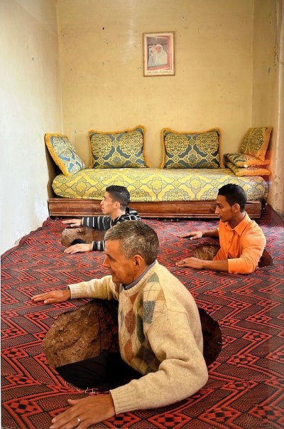 A photograph of three men appearing to emerge from holes cut into the carpeted floor of a living room.