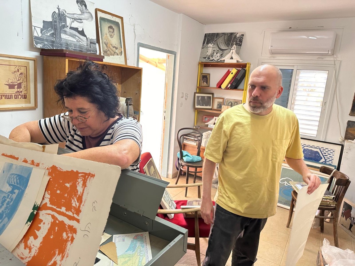 A bald man with a beard in a yellow shirt and a woman with dark hair and glasses in a striped shirt look at various prints.