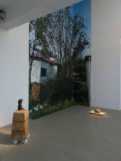 A large photograph of a tree between two houses pasted to the corner of a wall. Two sculptures lie on the floor before it.