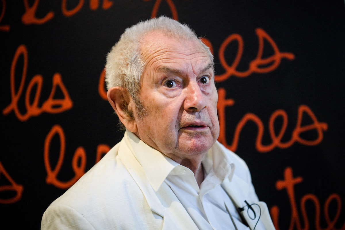 A smirking white man in a white shirt standing before a large painting composed of maroon text against a black background.