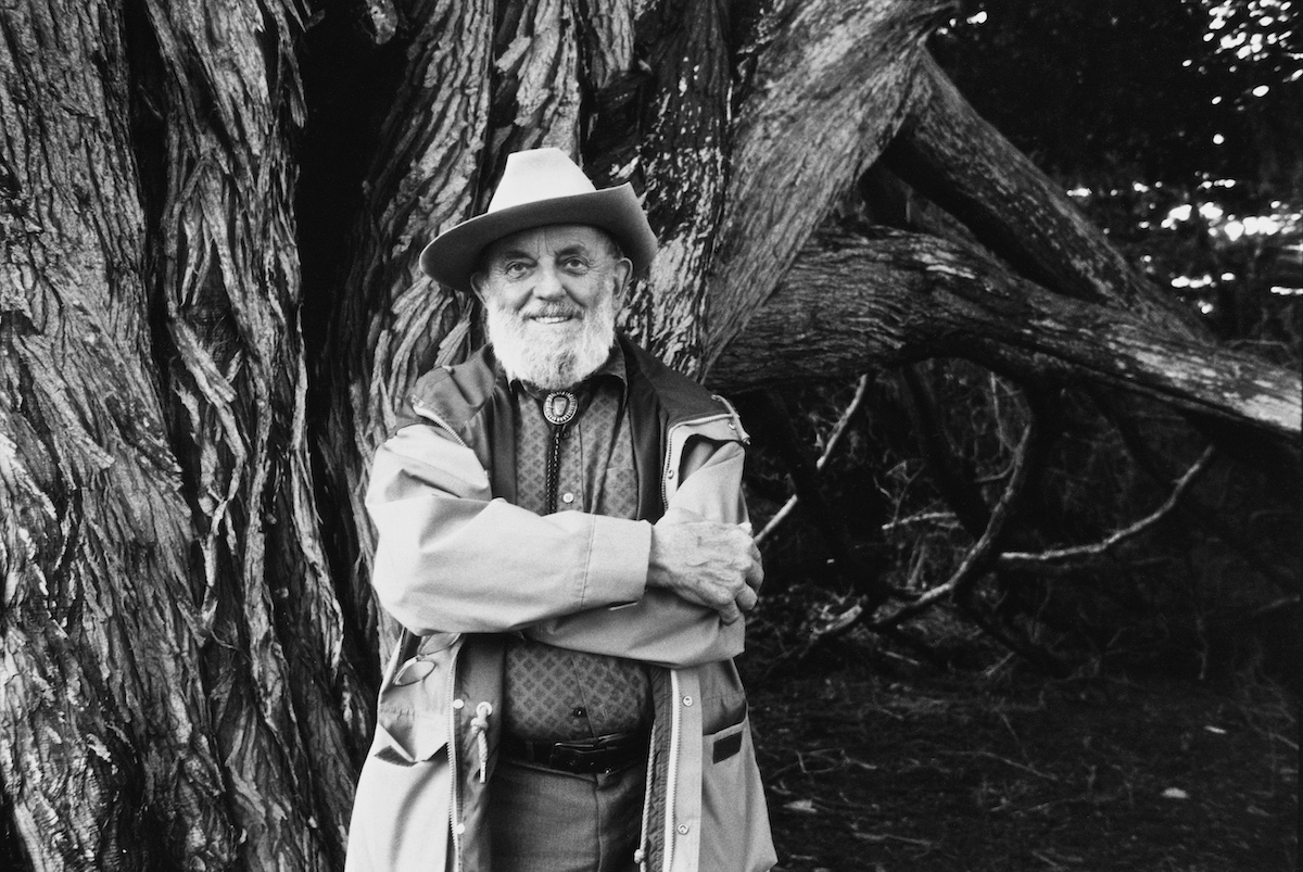 A white man in a hat crossing his arms and smiling in front of a tall tree.