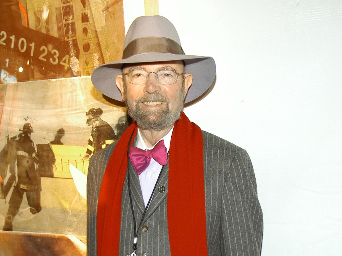 A smiling white man with a goatee in a grey brimmed hat. He wears a red scarf around a pinstriped suit and a pink bow tie. He stands in front of a large reproduction of a black-and-white photograph.