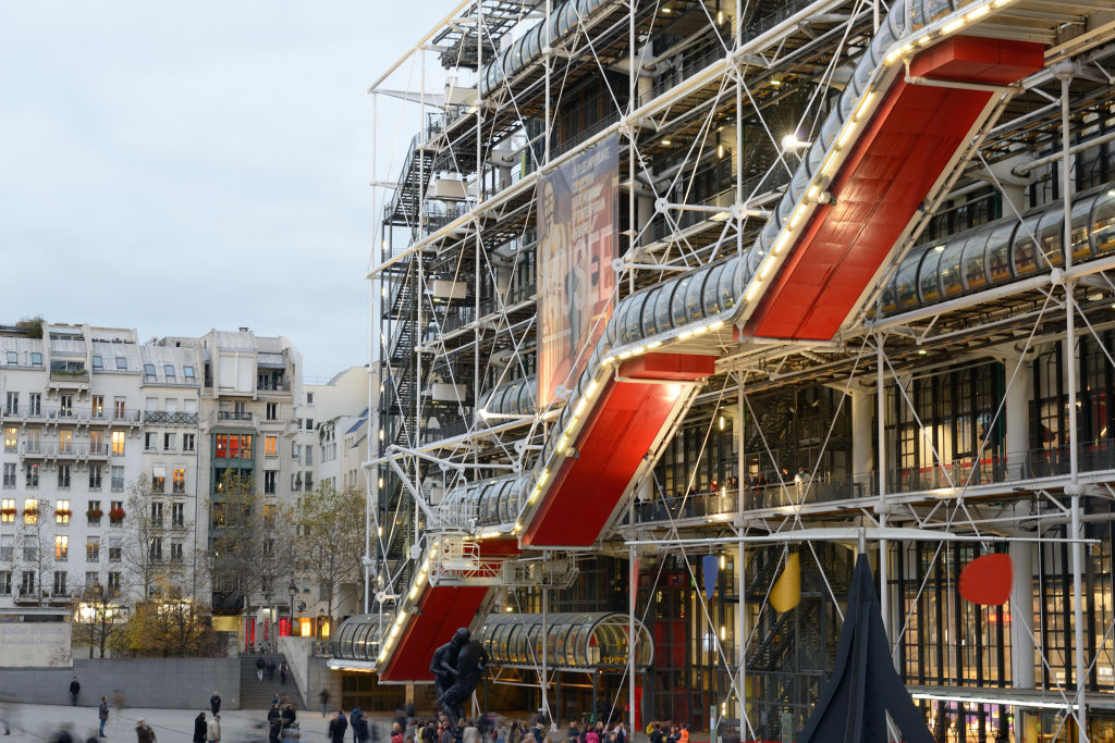 An angular building with piping on its exterior with people beneath.