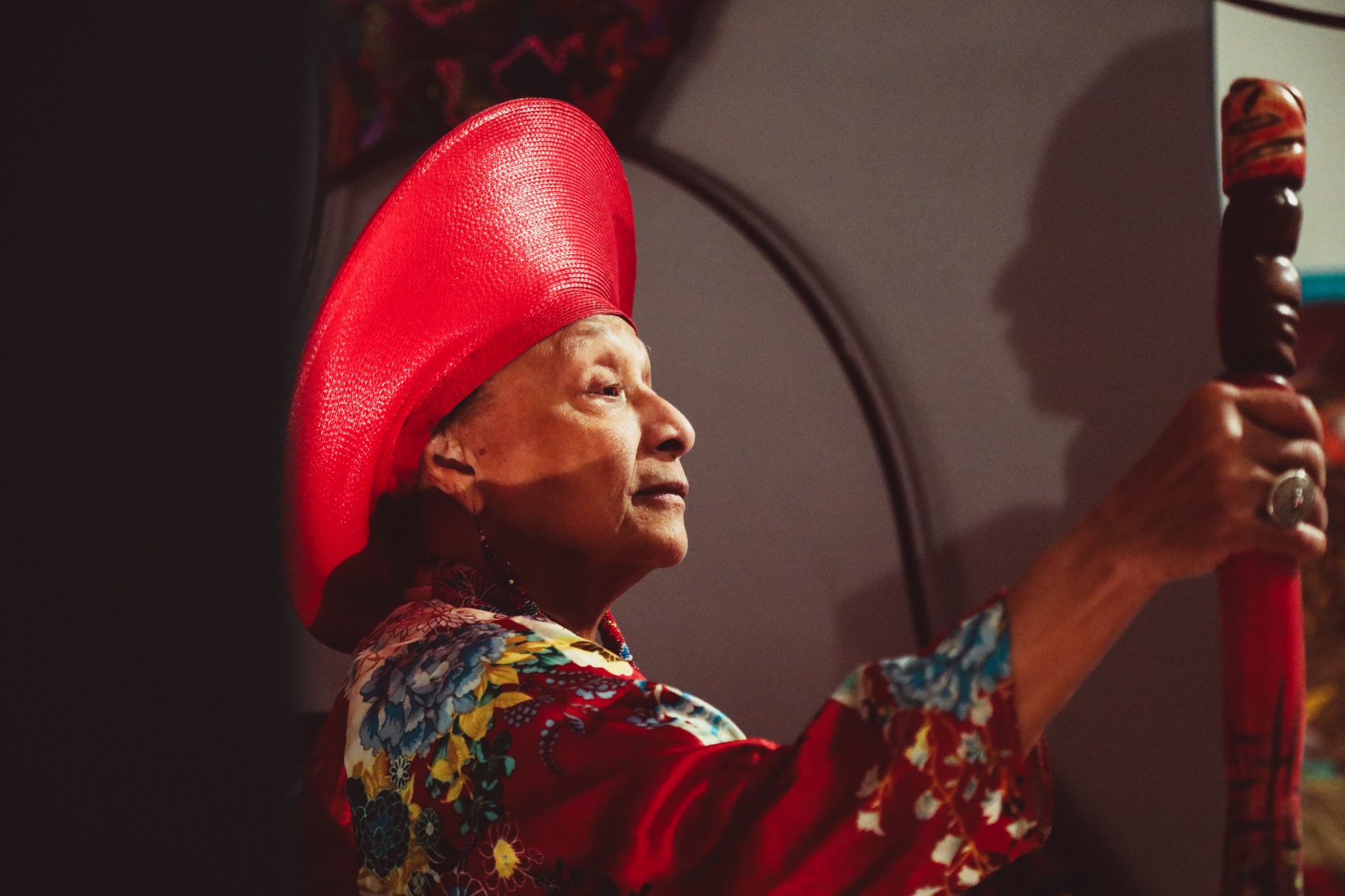 A Black woman in profile wearing a big red hat and holding a wooden staff.