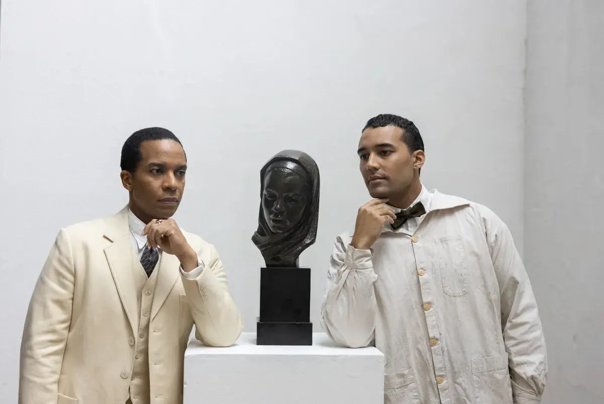 Two brown-skinned men stand on either side of a pedestal with a dark sculptural bust between them. They wear light colored clothing and each rests an elbow on the pedestal as they gaze at the sculpture.