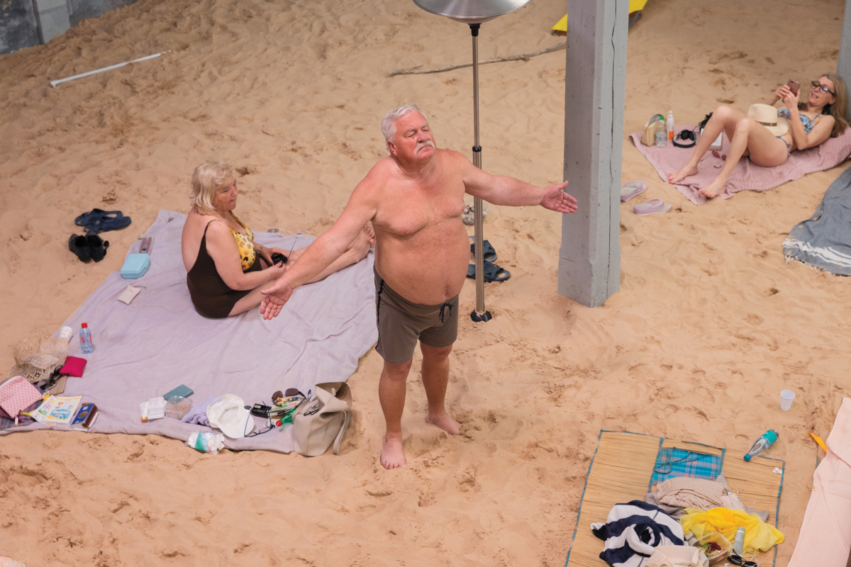 A blubbery shirtless white man stands with arms outstretched on a fake beach as people lounge on towels in the sad.